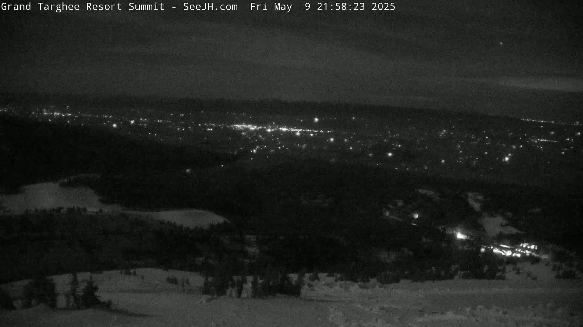 A nighttime, snowy, high-angle view shows a distant city illuminated by lights below a dark, treeline.