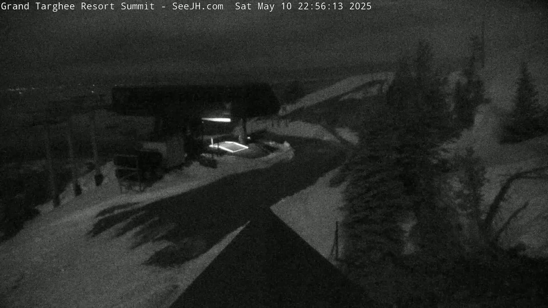 A nighttime, snow-covered scene shows a building and a road, surrounded by dark evergreen trees under a dark sky.