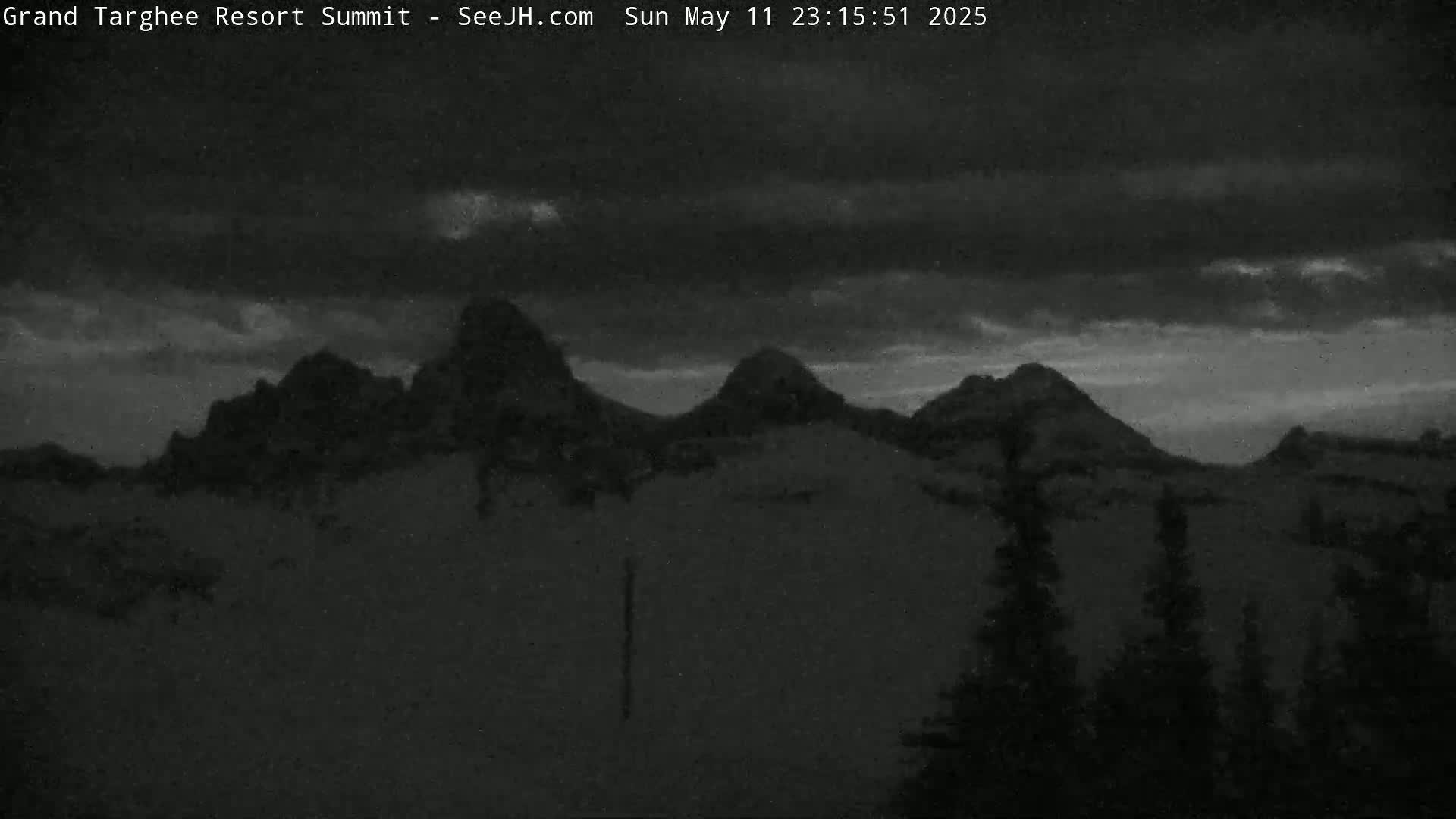 A dark, cloudy night scene shows a range of silhouetted mountains, with a few dark trees in the foreground.