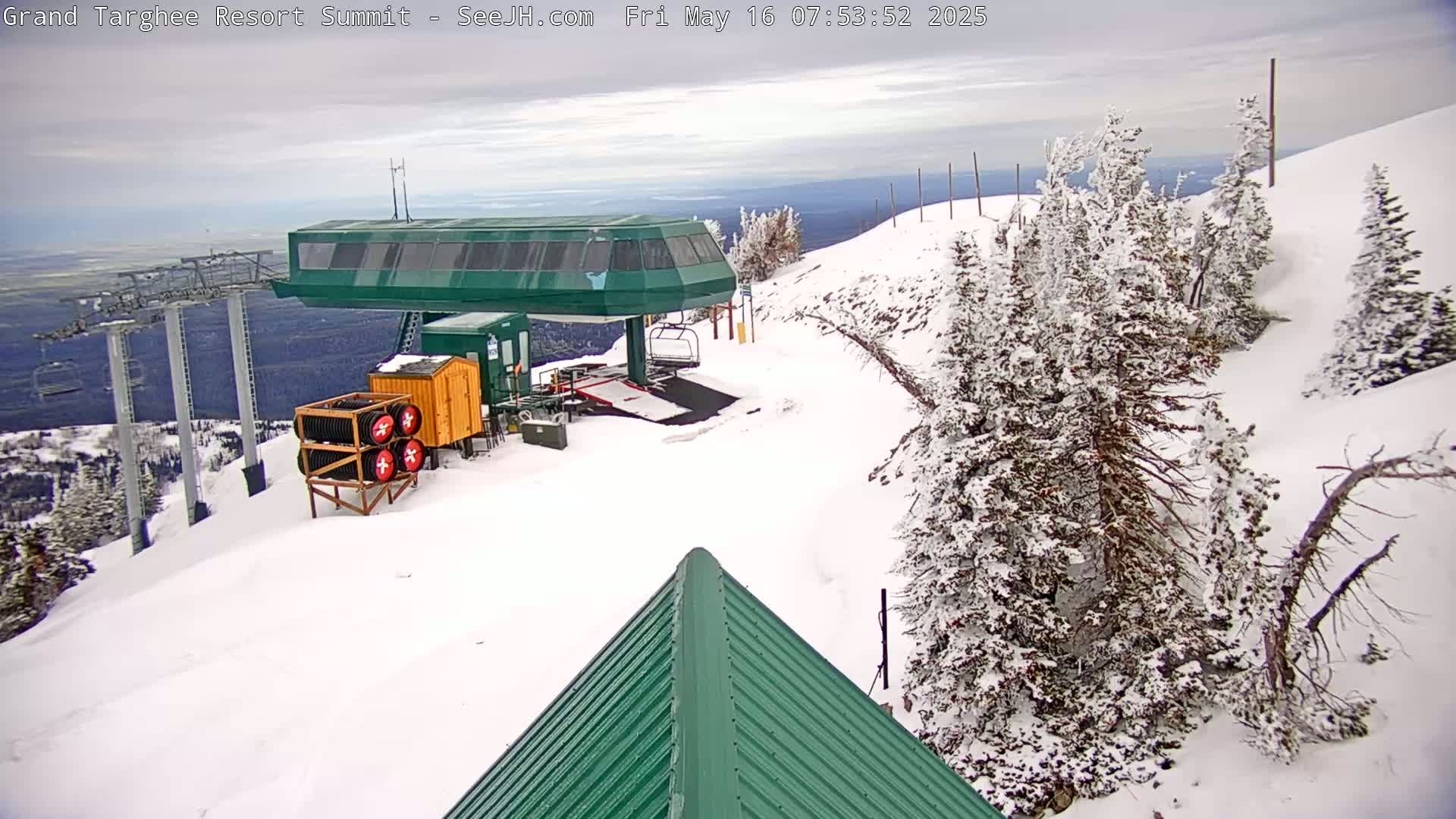 A snow-covered mountaintop ski lift station with overcast skies and snow-laden evergreens.