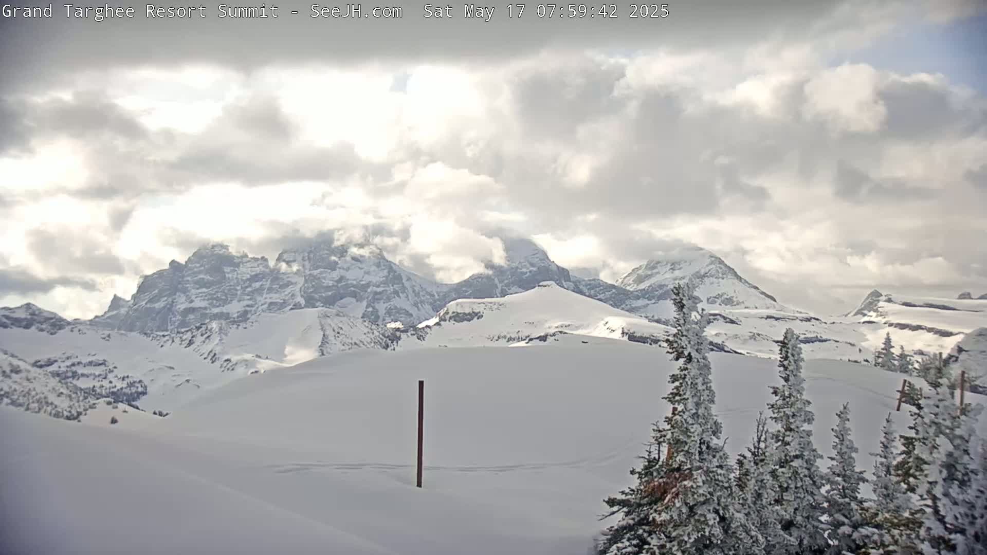 A snow-covered mountain range is visible under a partly cloudy sky, with snow-covered evergreens in the foreground.