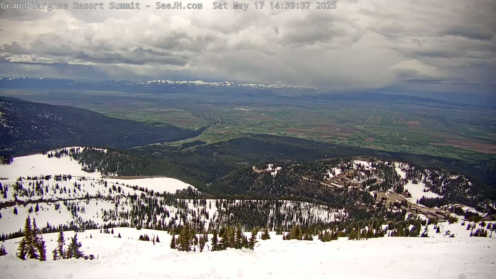 A snow-covered mountain peak overlooks a valley of green fields and distant, snow-capped mountains under a cloudy sky.