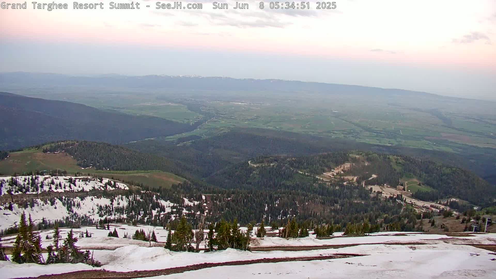 A hazy, sunrise view from a snow-covered mountaintop shows a vast valley with green fields and distant mountains.