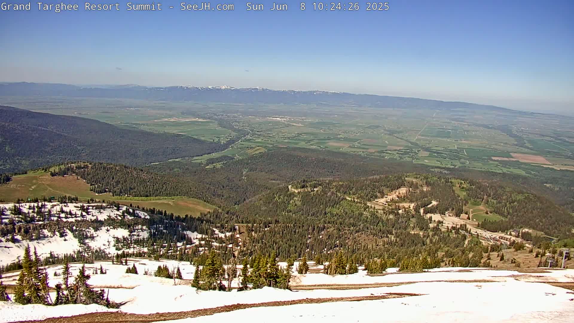 A high-altitude view on a clear, sunny day shows a vast valley of green farmland stretching to distant mountains, with a closer view of a snow-dusted mountainside dotted with evergreens.
