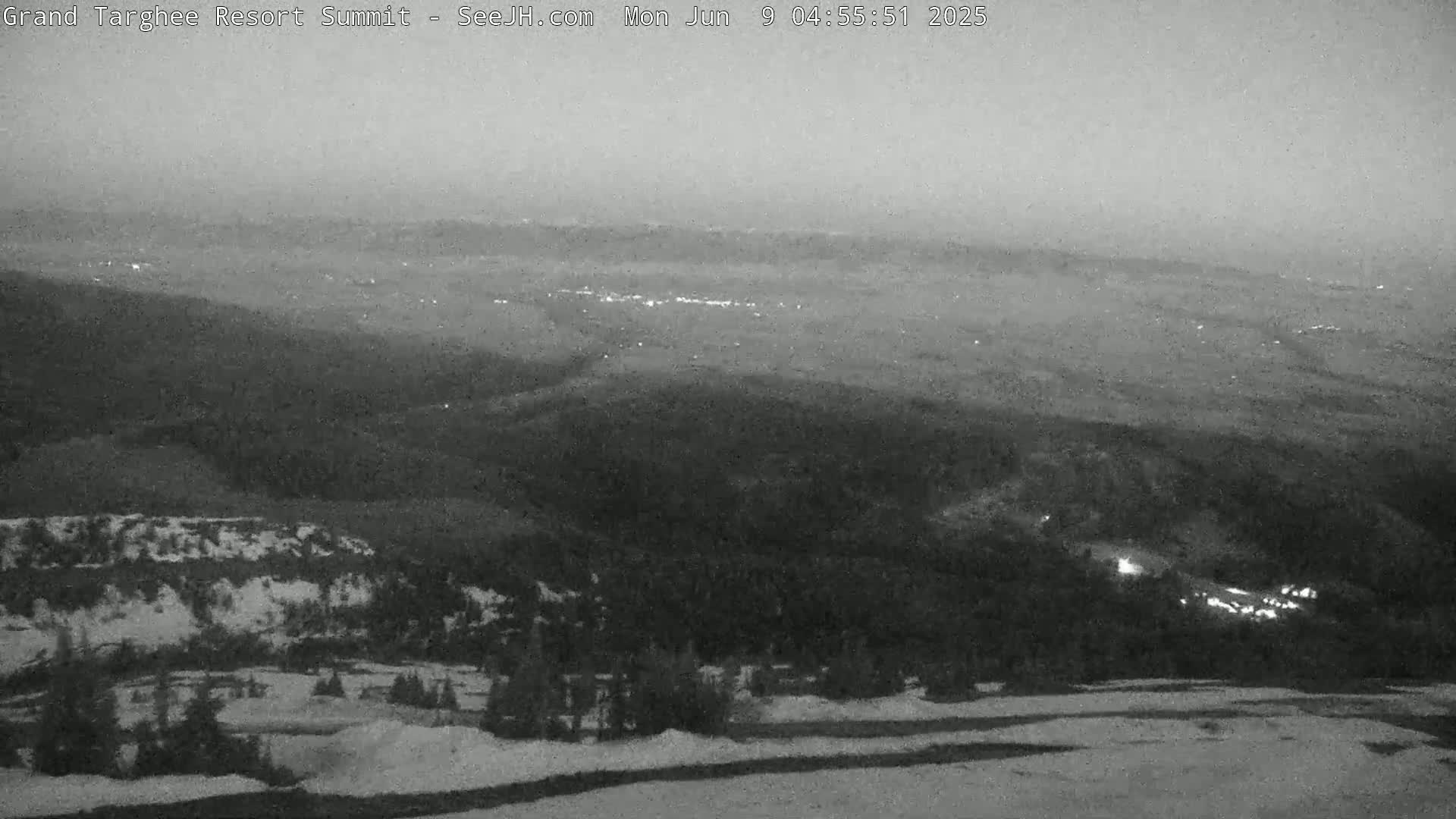 A hazy, pre-dawn view of a mountainous landscape shows patches of snow and distant lights in a valley below.