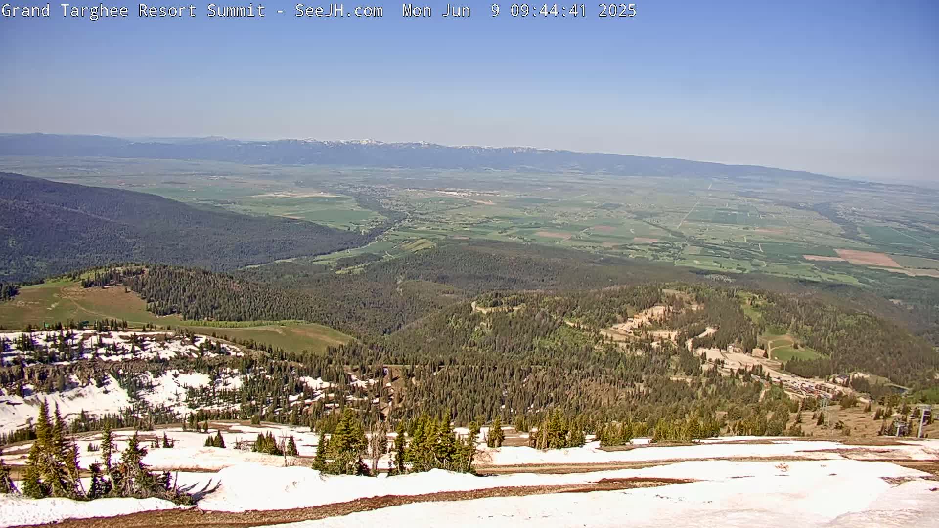 A high-altitude view on a clear, sunny day shows a vast expanse of green valley and distant mountains, with patches of snow remaining on the nearby slopes of a forested mountain.