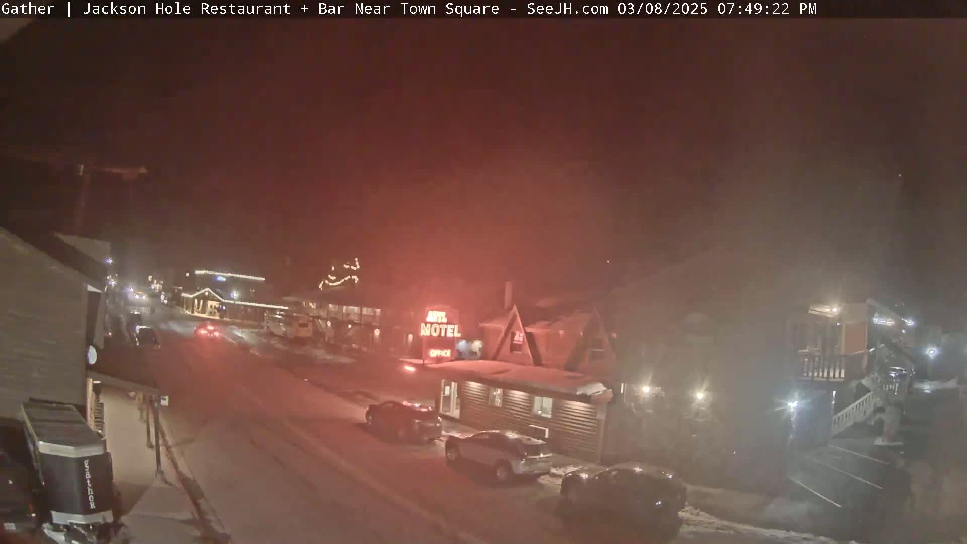 A snow-covered street at night, dimly lit with vehicles parked and driving past buildings including a motel.