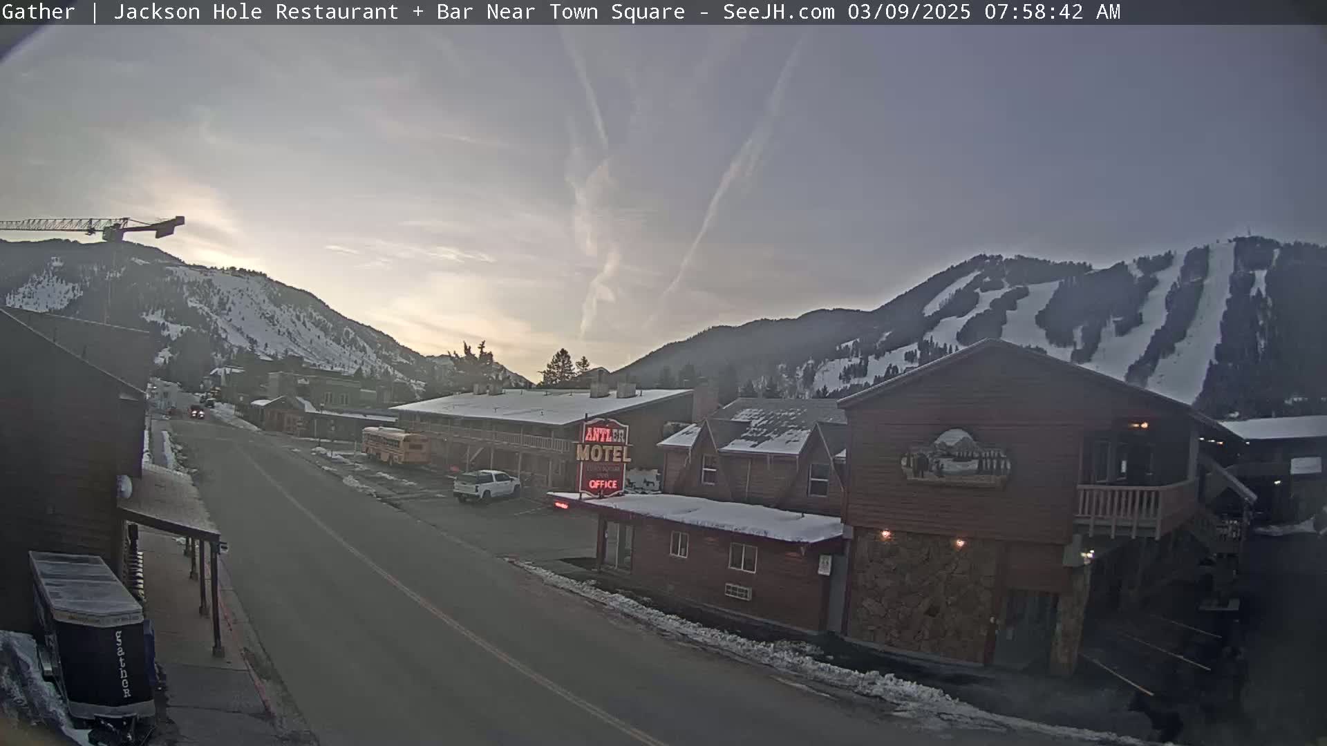 A mostly clear, dusky morning reveals a snow-covered mountainside with ski slopes behind a street scene featuring a motel, buildings, vehicles, and a construction crane.