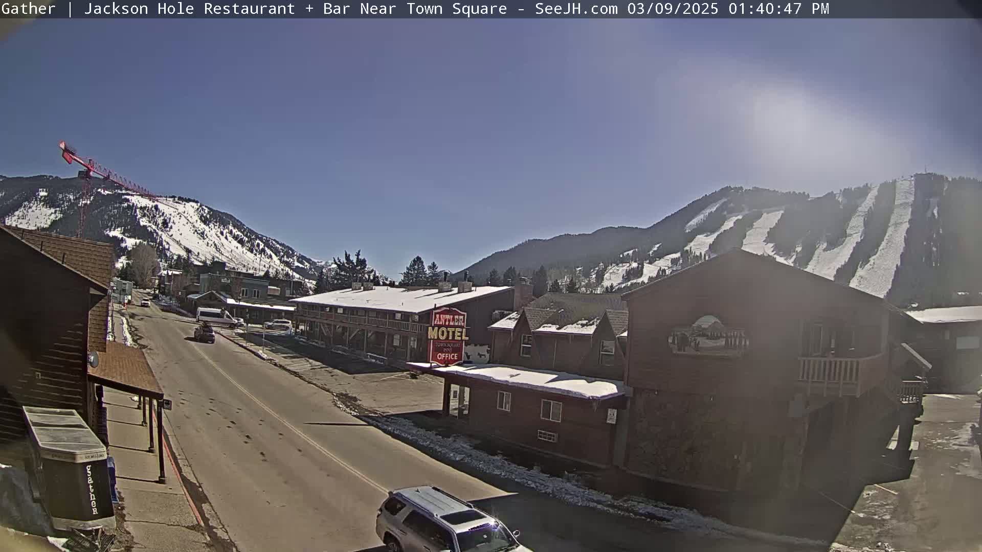 A sunny day reveals a snow-covered mountainside with ski runs, overlooking a town street with buildings, cars, and a motel.