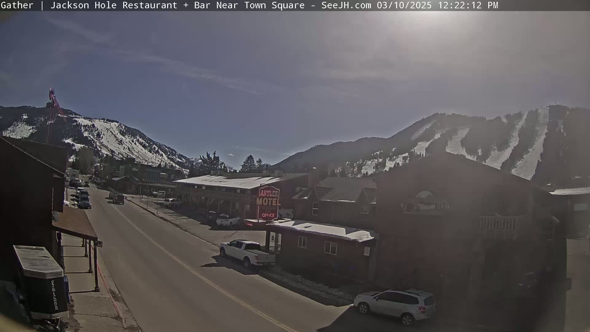 A sunny day reveals a snow-dusted mountain range overlooking a town street with parked cars and buildings, including a motel.