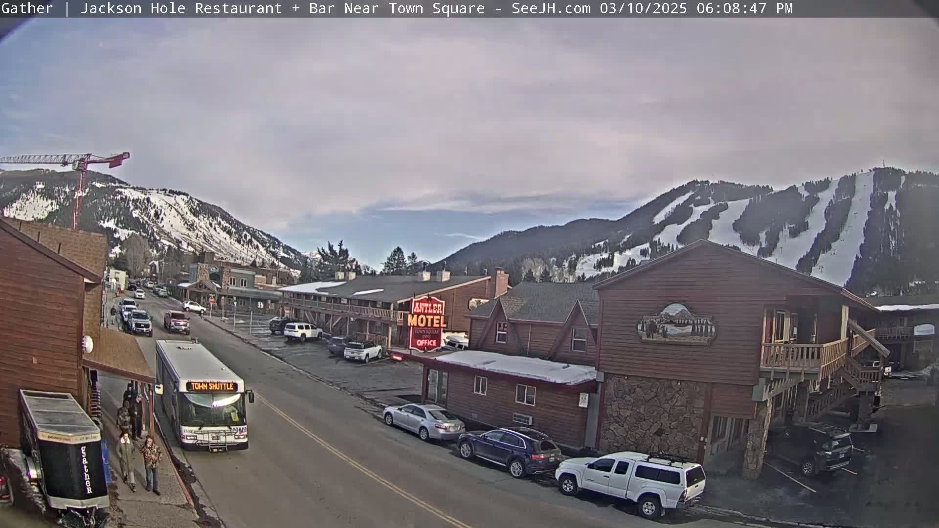 A snow-covered mountainside overlooks a town street with cars parked along the roadside, a town shuttle bus stopped at a curb, and several buildings including a motel.