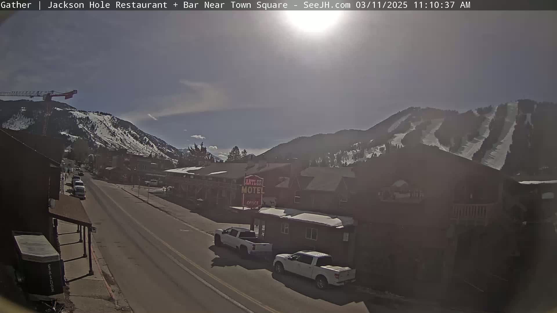 A sunny day reveals a street scene in a mountain town with snow-covered hills in the background, several cars parked along the road, and buildings lining the street.