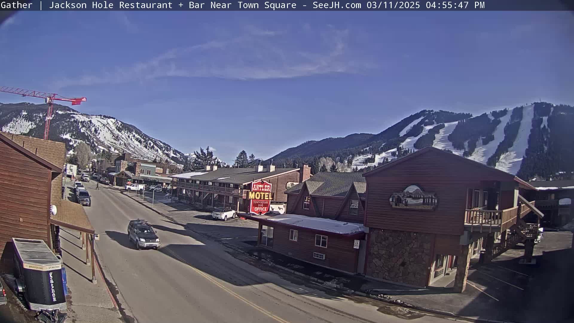 A sunny day reveals a street scene in a mountain town with snow-covered slopes in the background, vehicles parked along the roadside, and a motel in the foreground.