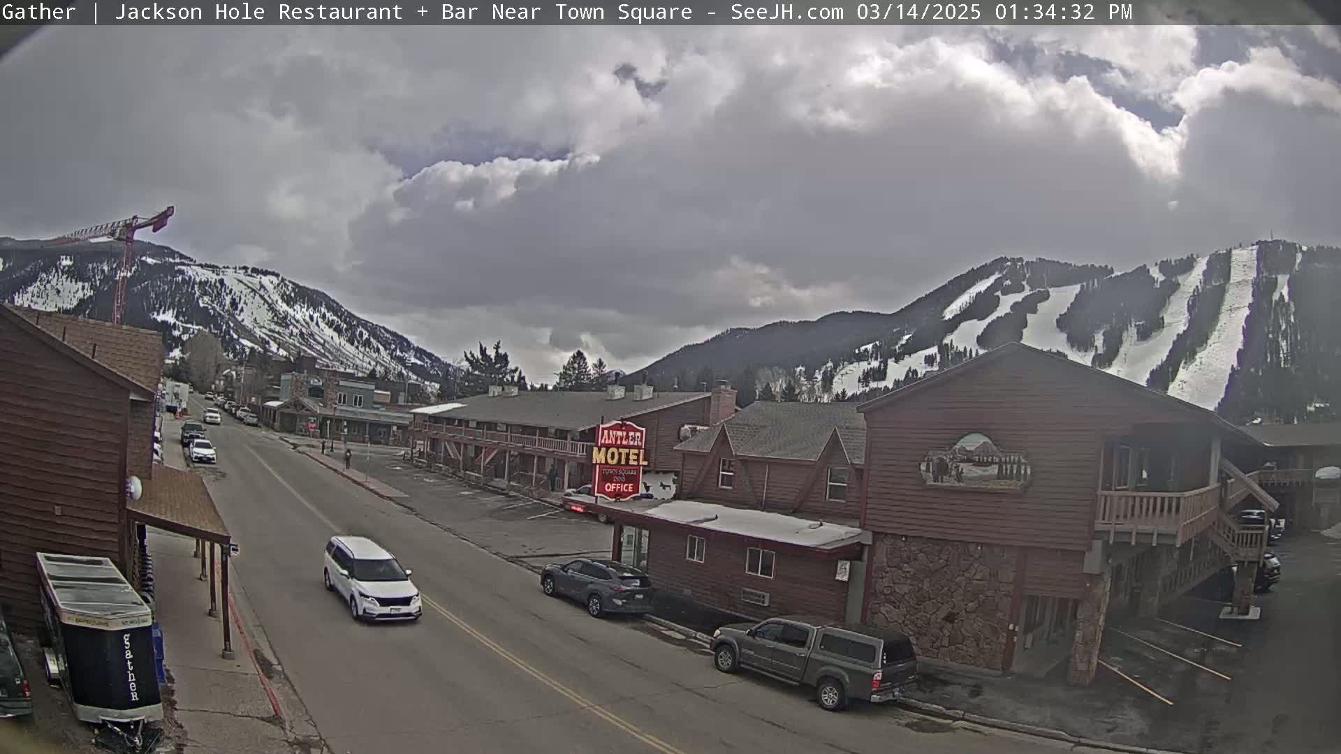 A partly cloudy day reveals a snow-covered mountainside with ski runs in the background, overlooking a street scene with cars and buildings, including a motel.
