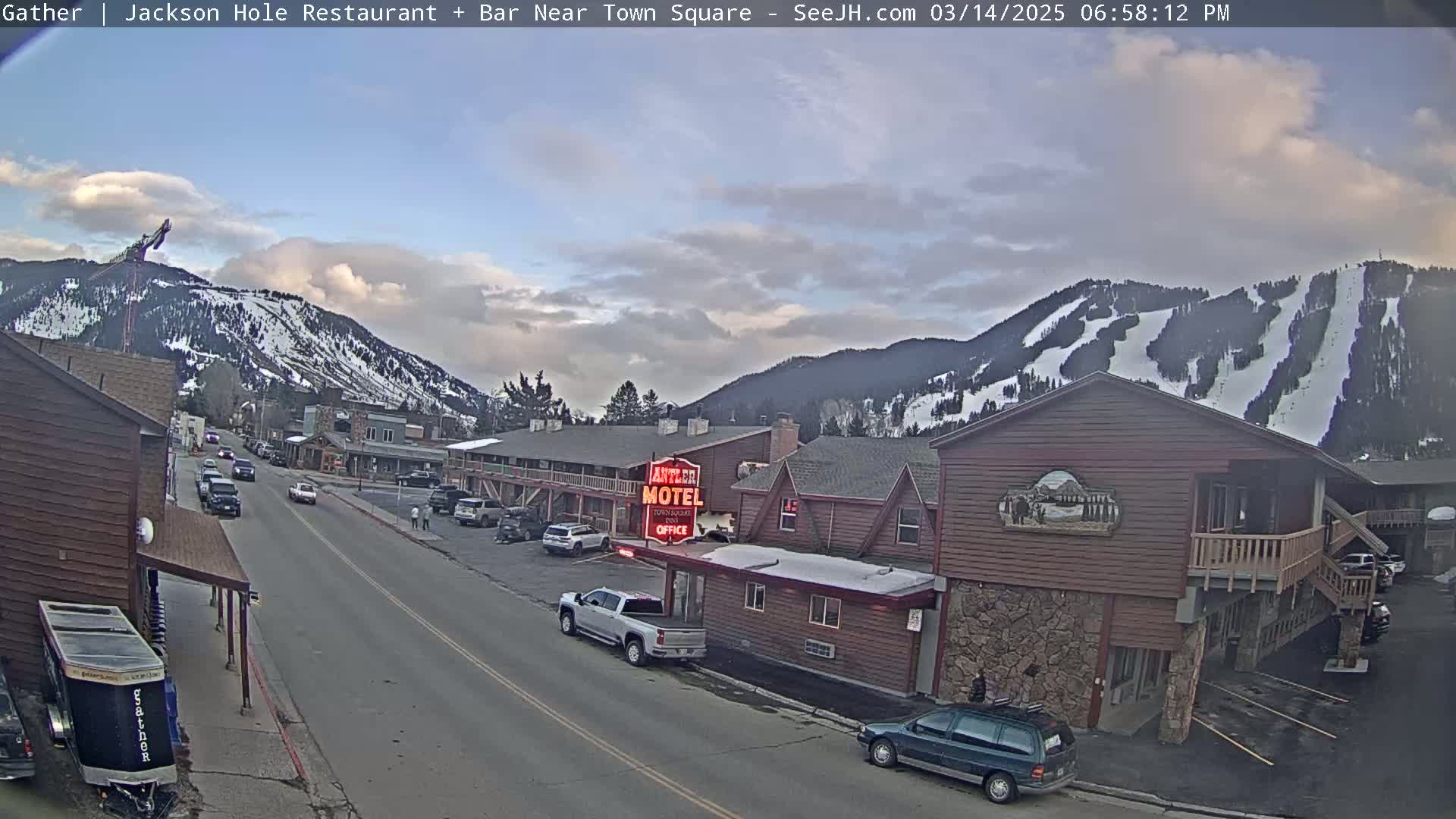 A partly cloudy afternoon scene shows a street in a town nestled in a valley with snow-covered mountains and ski slopes in the background.