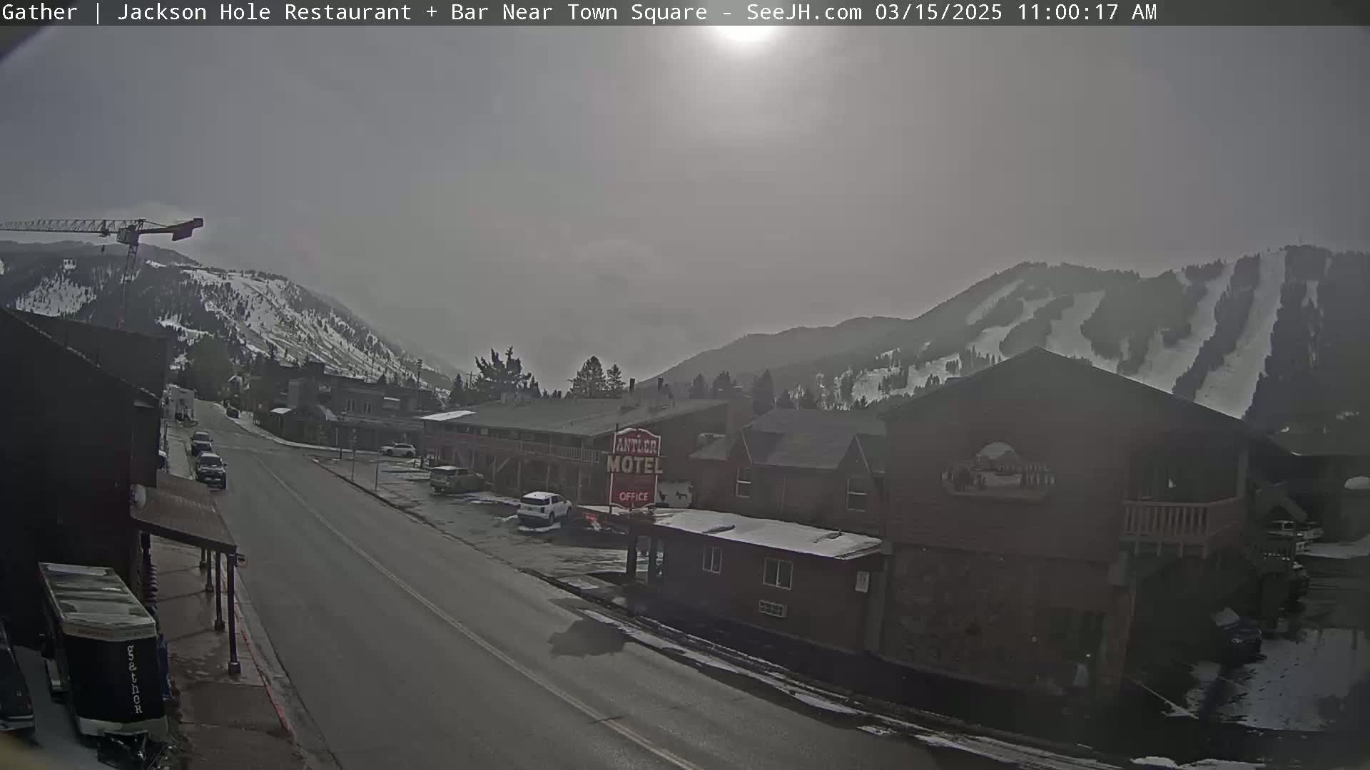 A hazy, overcast day reveals a street scene in a town nestled among snow-capped mountains, with a motel and some vehicles visible.