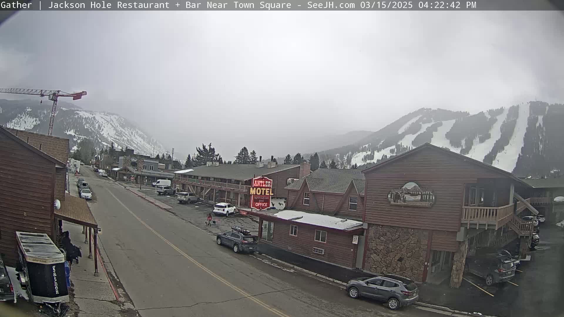 A mostly cloudy day reveals a street scene with cars parked alongside buildings, a motel, and a crane in the background, all set against snow-covered mountains.