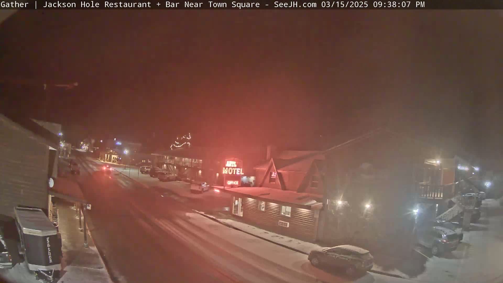 A snow-covered street at night, showing a motel and other buildings with cars parked along the roadside under a hazy sky.