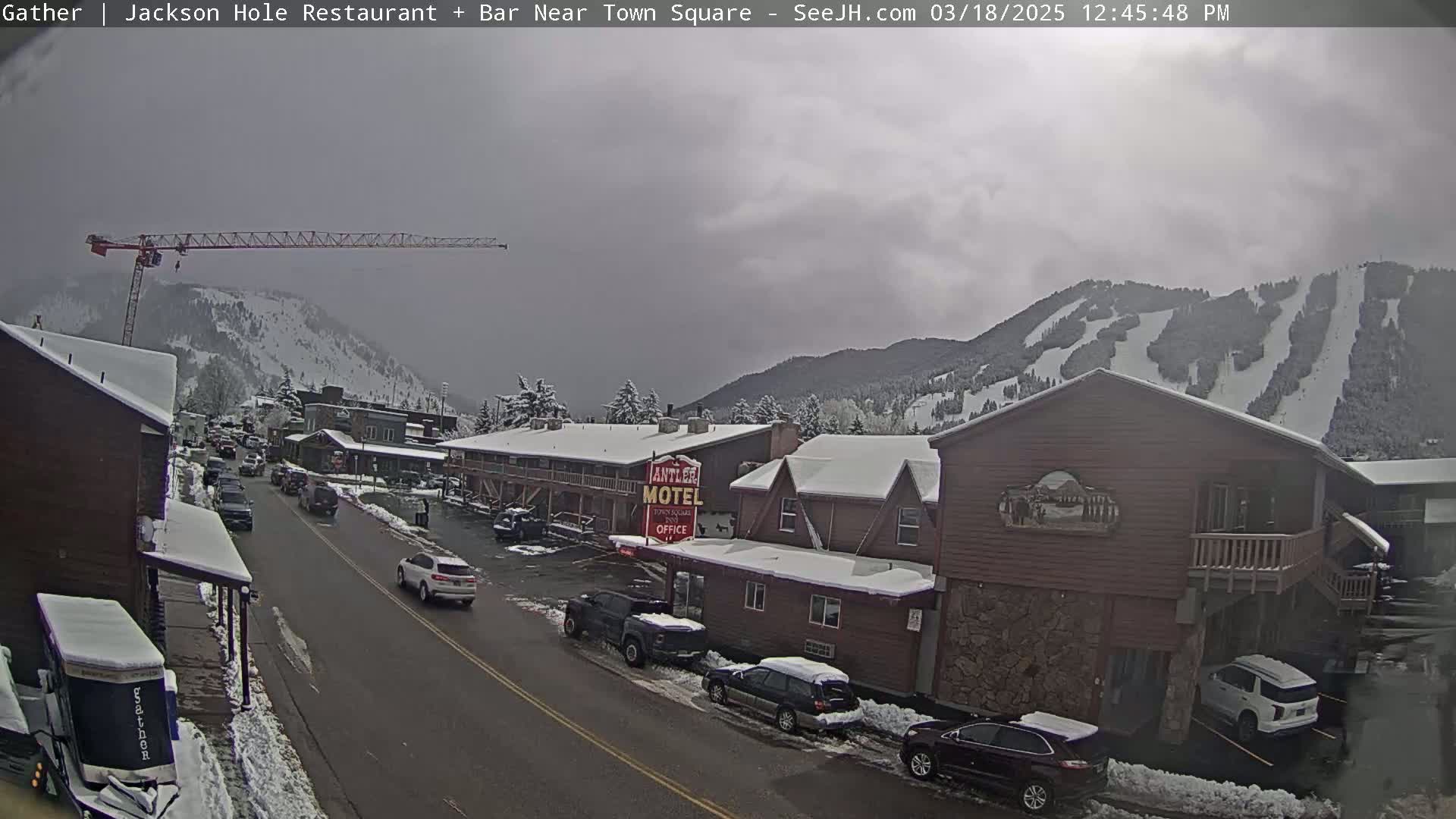 A snow-covered town street scene with cars parked along the sides, a motel, and a construction crane visible against a backdrop of snow-covered mountains under an overcast sky.