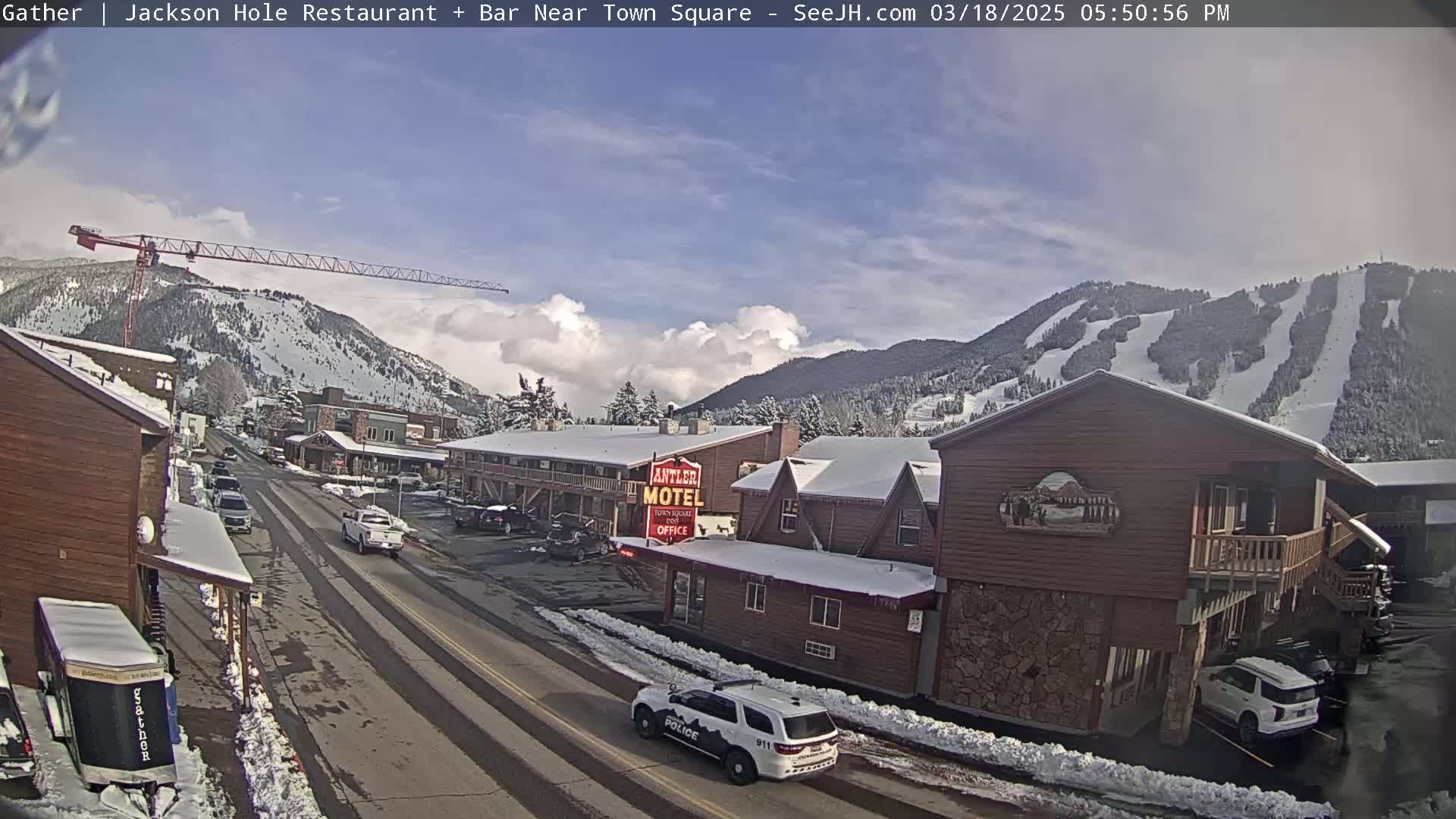 A partly cloudy day reveals a snow-covered town with a street, buildings, a motel, and a construction crane, all nestled against a mountain with ski runs.