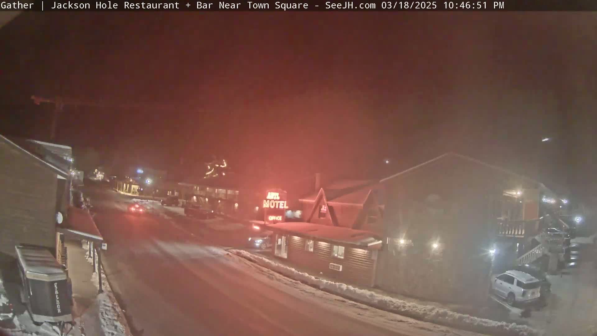 A nighttime street scene shows a motel and other buildings lightly covered in snow under hazy conditions.