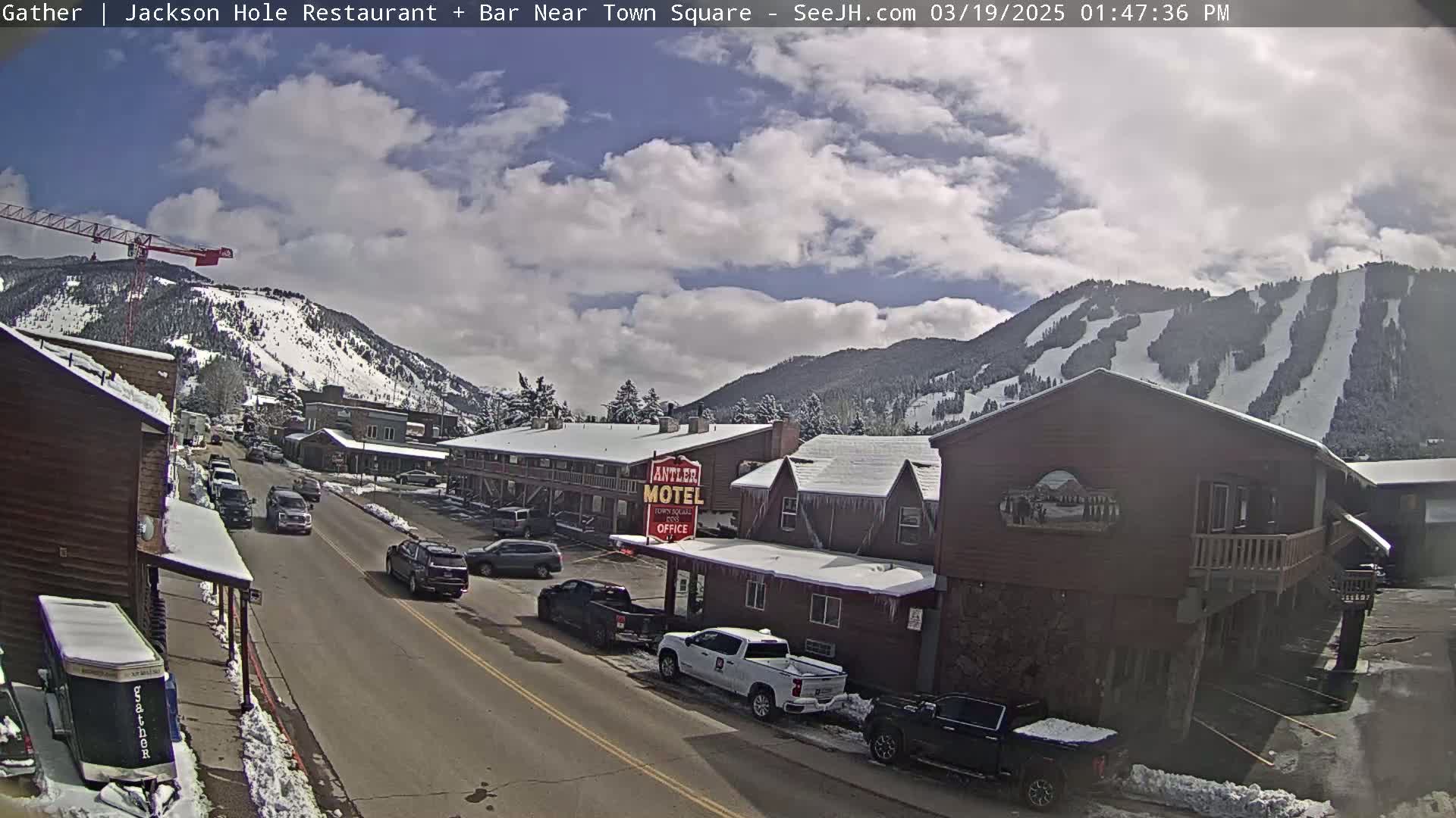 A partly cloudy day reveals a snow-dusted town with cars parked along a street, mountains in the background showing ski slopes, and a motel with icicles hanging from the roof.