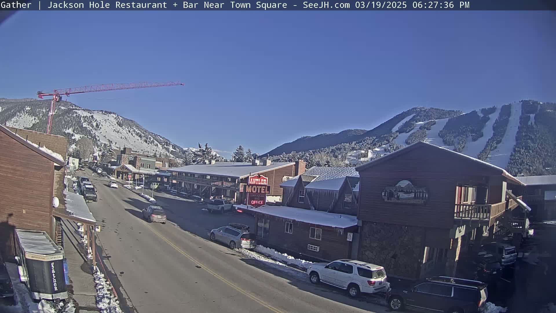 A sunny day reveals a street scene in a mountain town, with cars parked along the road, buildings with snow on their roofs, and a construction crane visible in the distance.