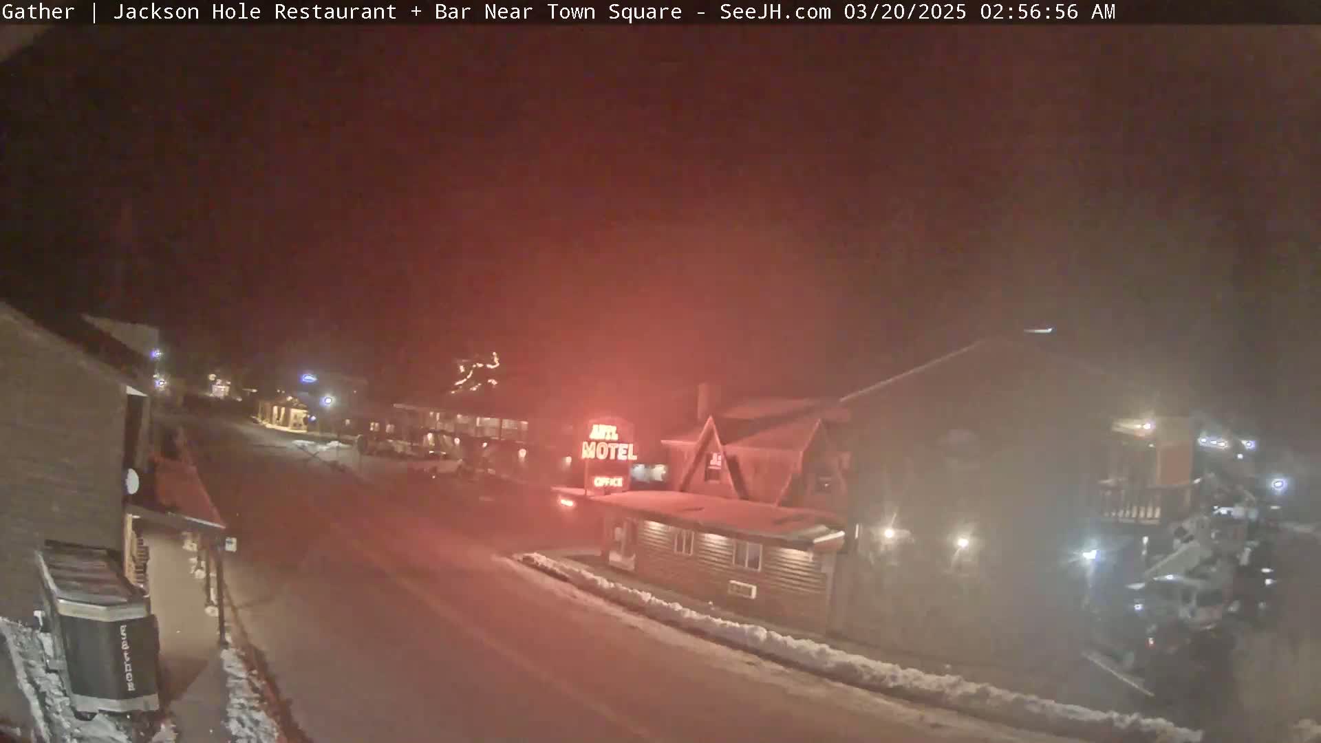 A nighttime, snowy street scene shows a motel with its sign illuminated, along with other buildings and vehicles under hazy conditions.