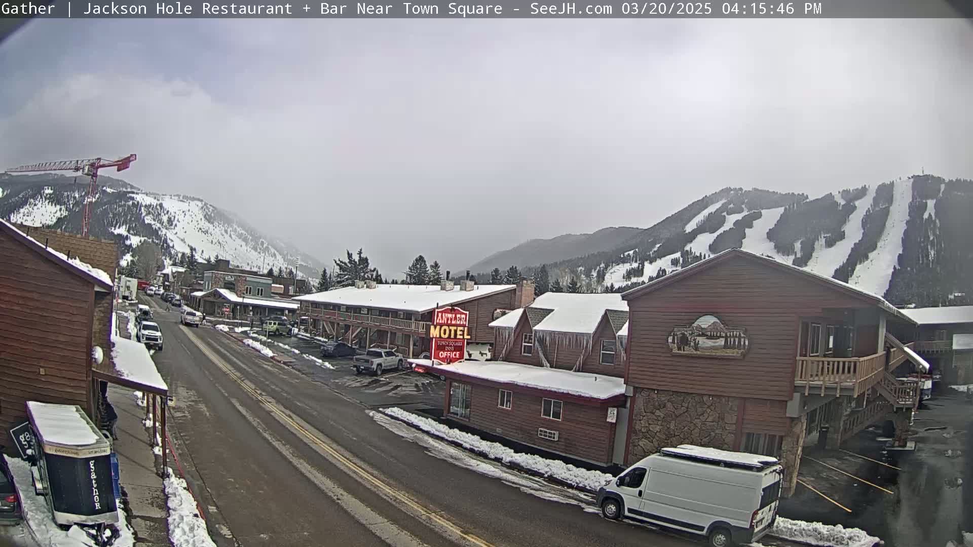 A snow-covered street scene shows a town nestled in a valley, with snow-covered mountains and ski slopes in the background, under an overcast sky.
