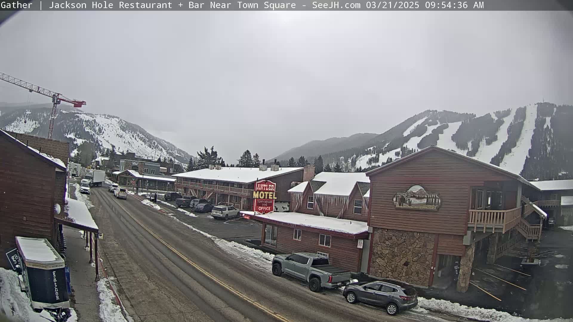 A snow-covered town street scene with cars parked along the sides, a motel, and snow-covered mountains in the background under an overcast sky.