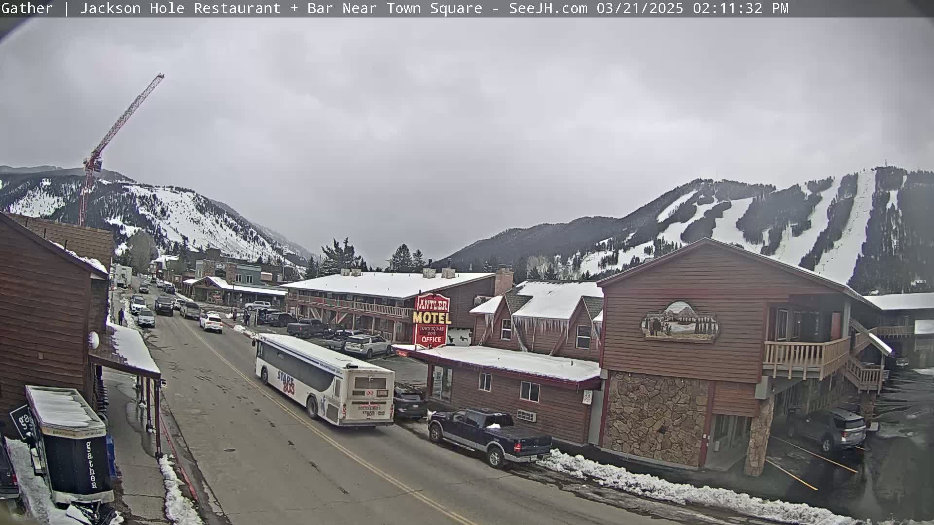 A street scene on an overcast day shows a town with snow-covered mountains in the background, a bus on the road, cars parked, and a motel.