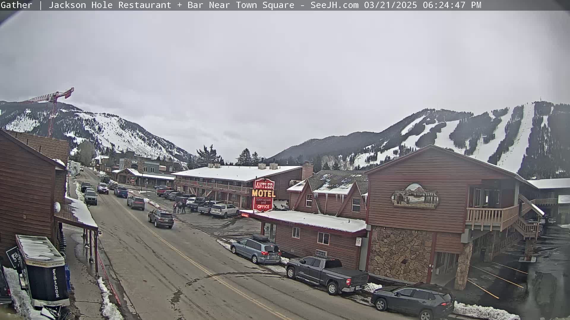 A street scene on an overcast day shows several vehicles parked along the sides of a road that runs past a motel and other buildings, with snow-covered mountains in the background.