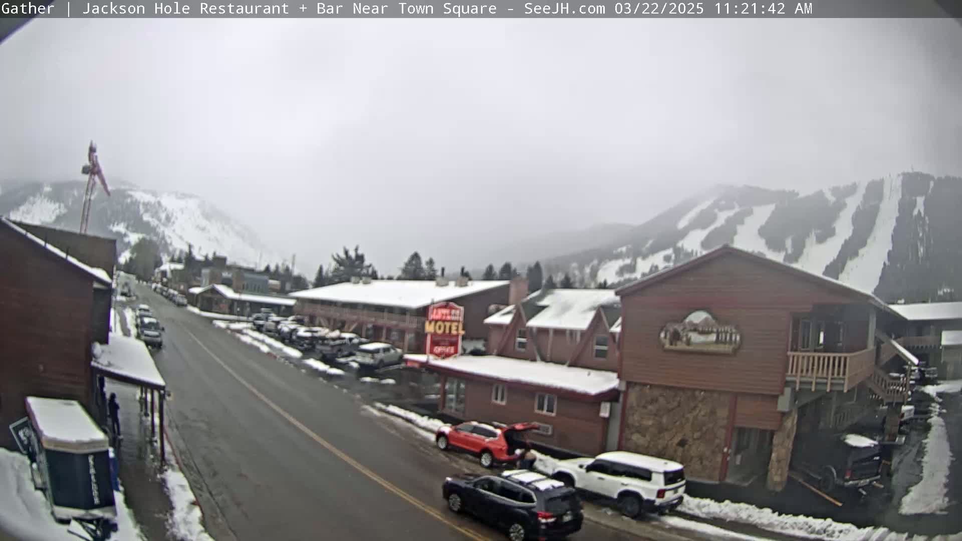 A snow-dusted street scene shows a town with parked cars, a motel, and snow-covered mountains in the background under an overcast sky.