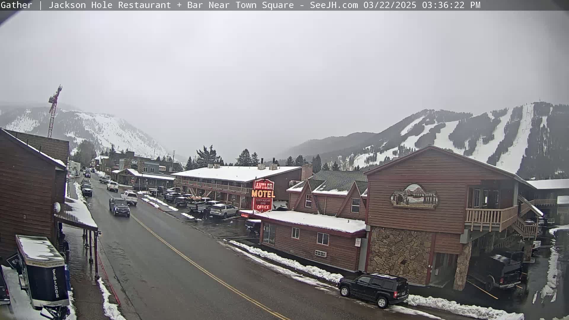 A snow-dusted town street scene with cars parked along the sides, several buildings including a motel, and snow-covered mountains in the background under an overcast sky.