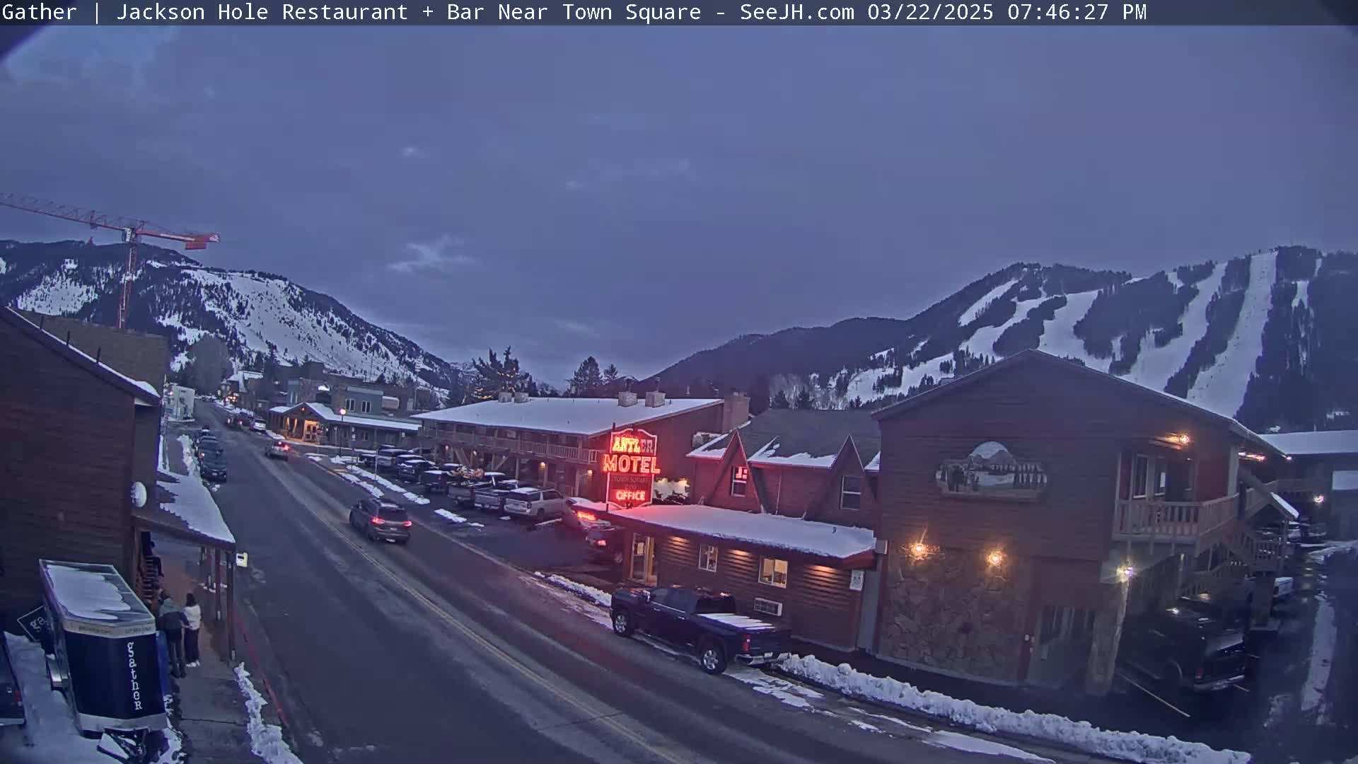 A twilight view of a snow-dusted town nestled in a valley beneath snow-covered mountains, with cars parked along a street next to a motel.