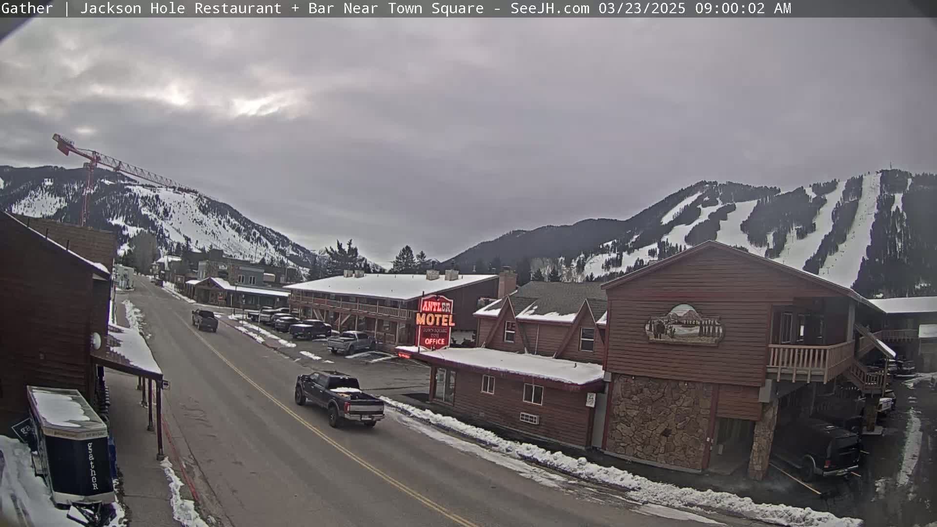 A street scene on an overcast day shows a snow-covered town with a motel, a construction crane, and snow-covered mountains in the background.