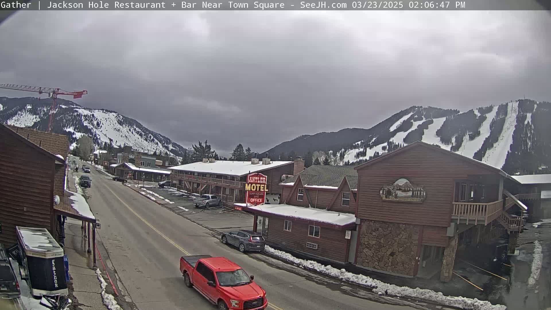 A street scene in a mountainous area under an overcast sky shows a road with cars and trucks, buildings including a motel, and snow on the ground and mountains in the background.