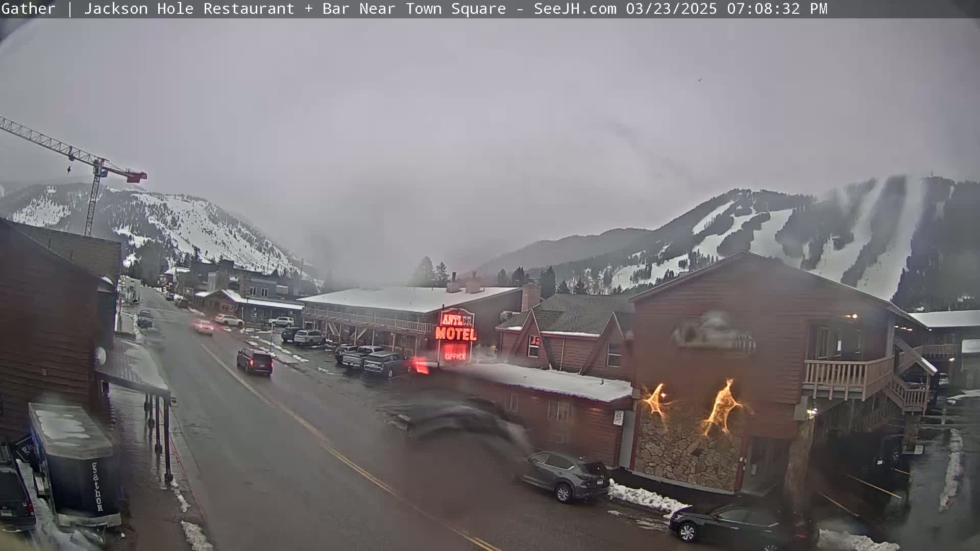 A wet, overcast day reveals a town street scene with cars parked and driving past a motel, a construction crane, and snow-covered mountains in the background.