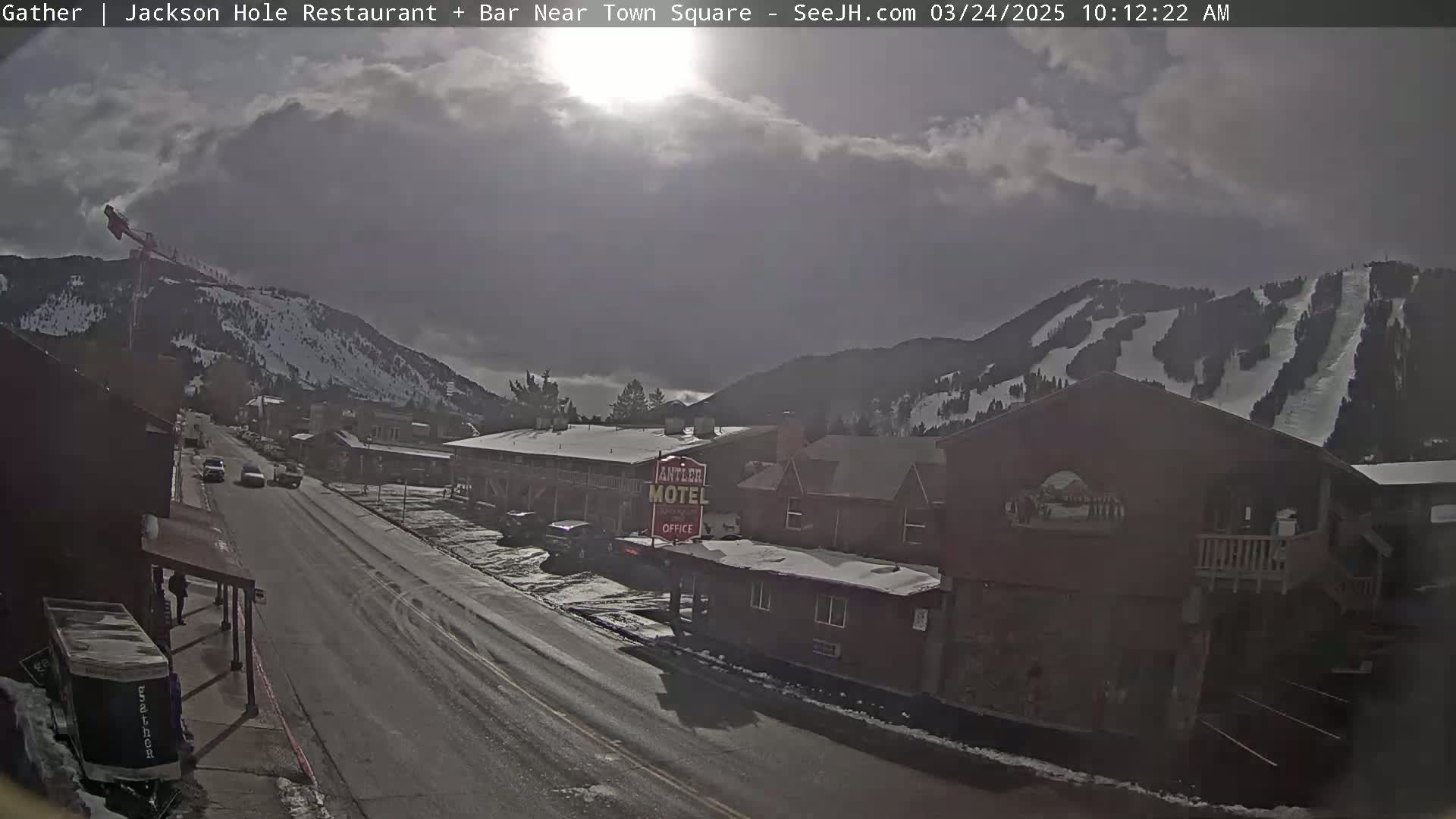 A partly cloudy day reveals a snow-covered mountain range overlooking a town street with buildings, cars, and a motel.