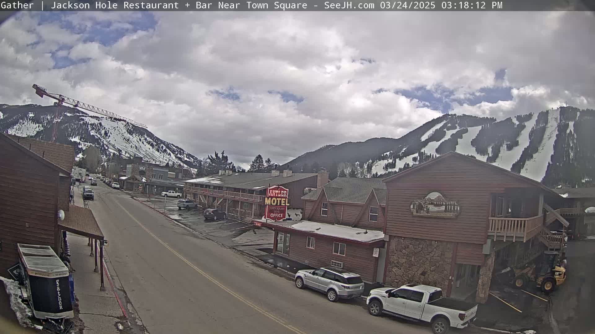 A partly cloudy day reveals a street scene in a mountain town, with snow-covered slopes visible in the background and parked cars lining the road in front of buildings.