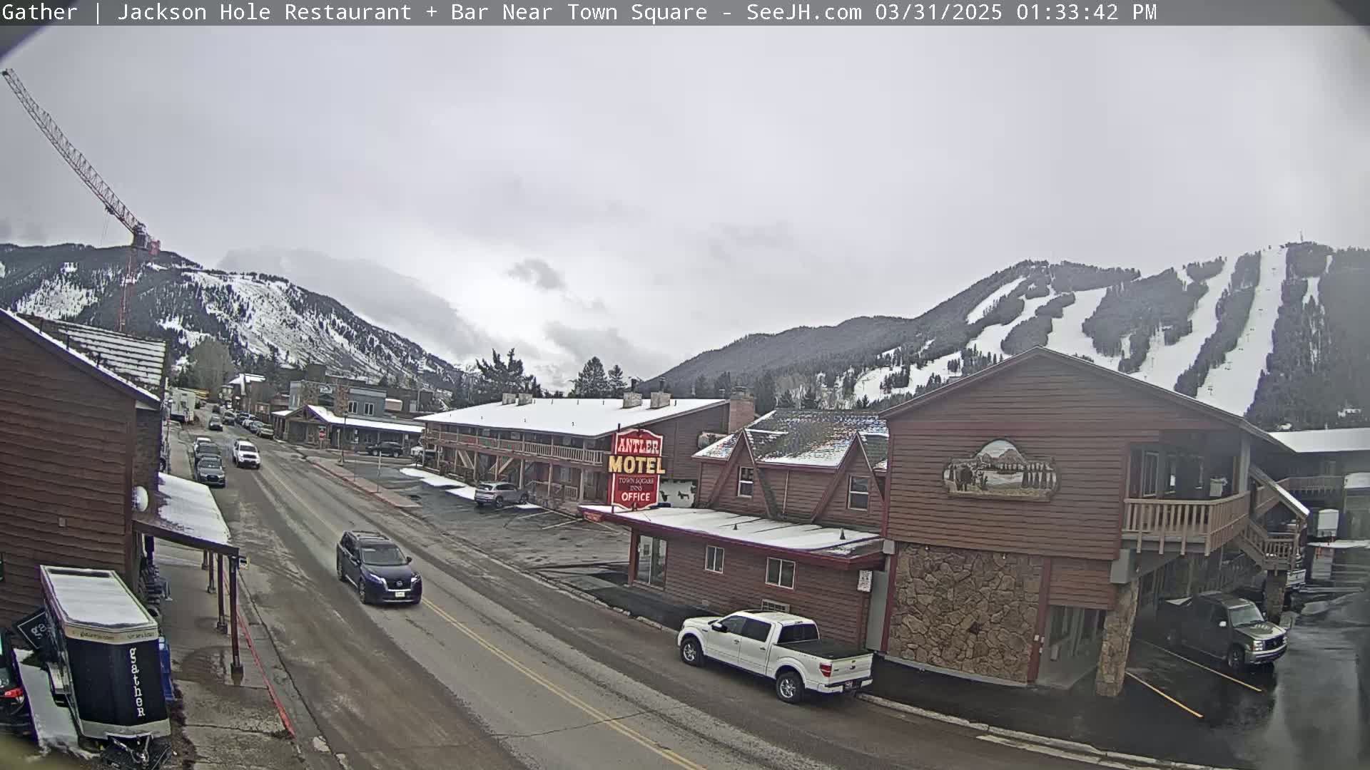 A snow-dusted town street scene unfolds under an overcast sky, with cars parked along the roadside and mountains showing ski runs in the background.