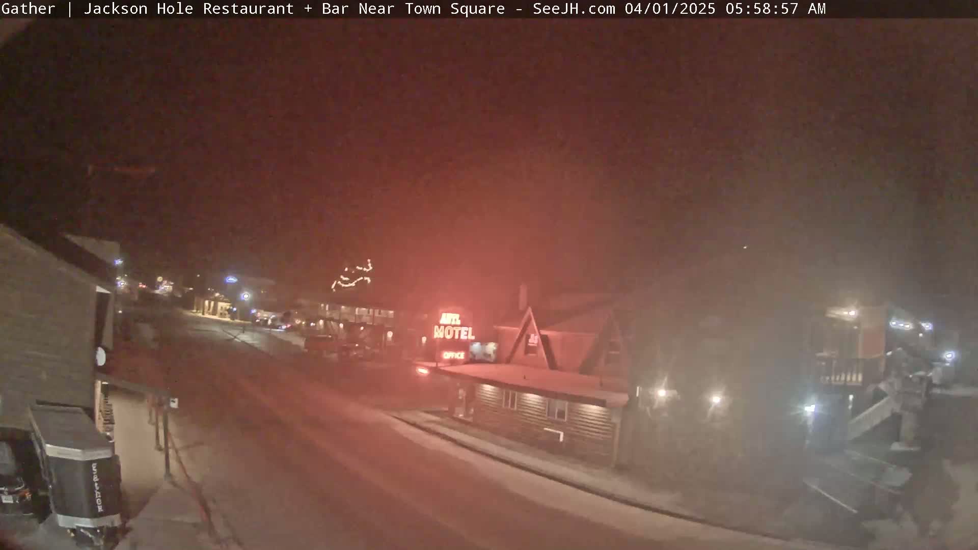 A snow-covered street at night, dimly lit with buildings and a motel visible in the distance under a hazy sky.