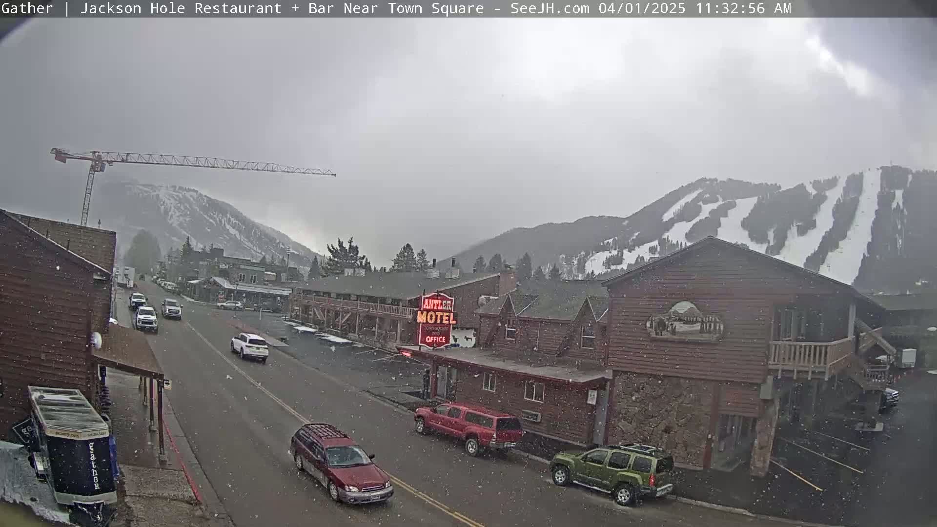 A lightly snowing scene shows a street with cars, a motel, and a construction crane, all set against a backdrop of snow-covered mountains with ski runs.