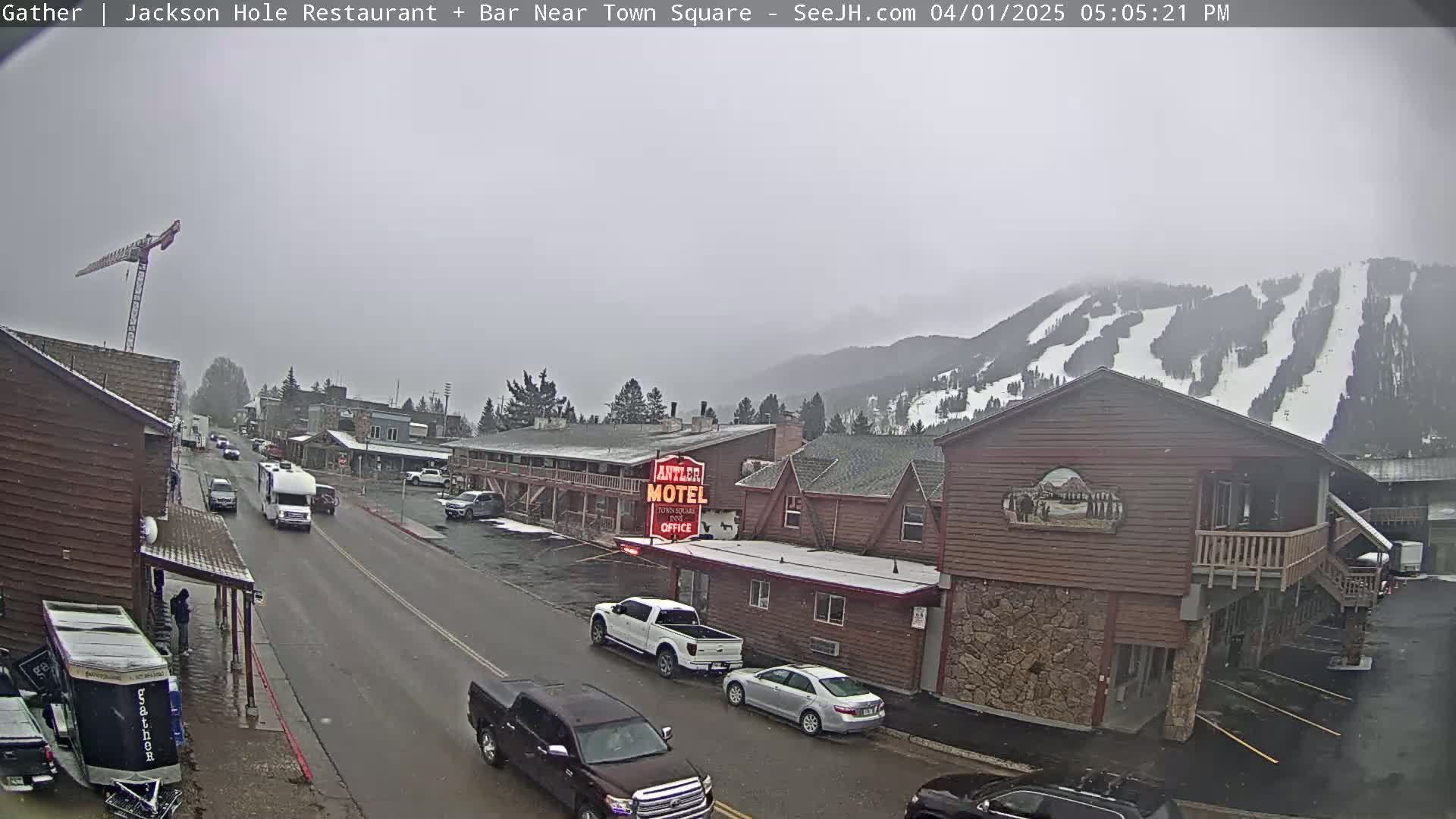 A street scene on an overcast day shows a motel and other buildings, with vehicles parked along the street and several cars driving, in front of snow-covered mountains in the background.