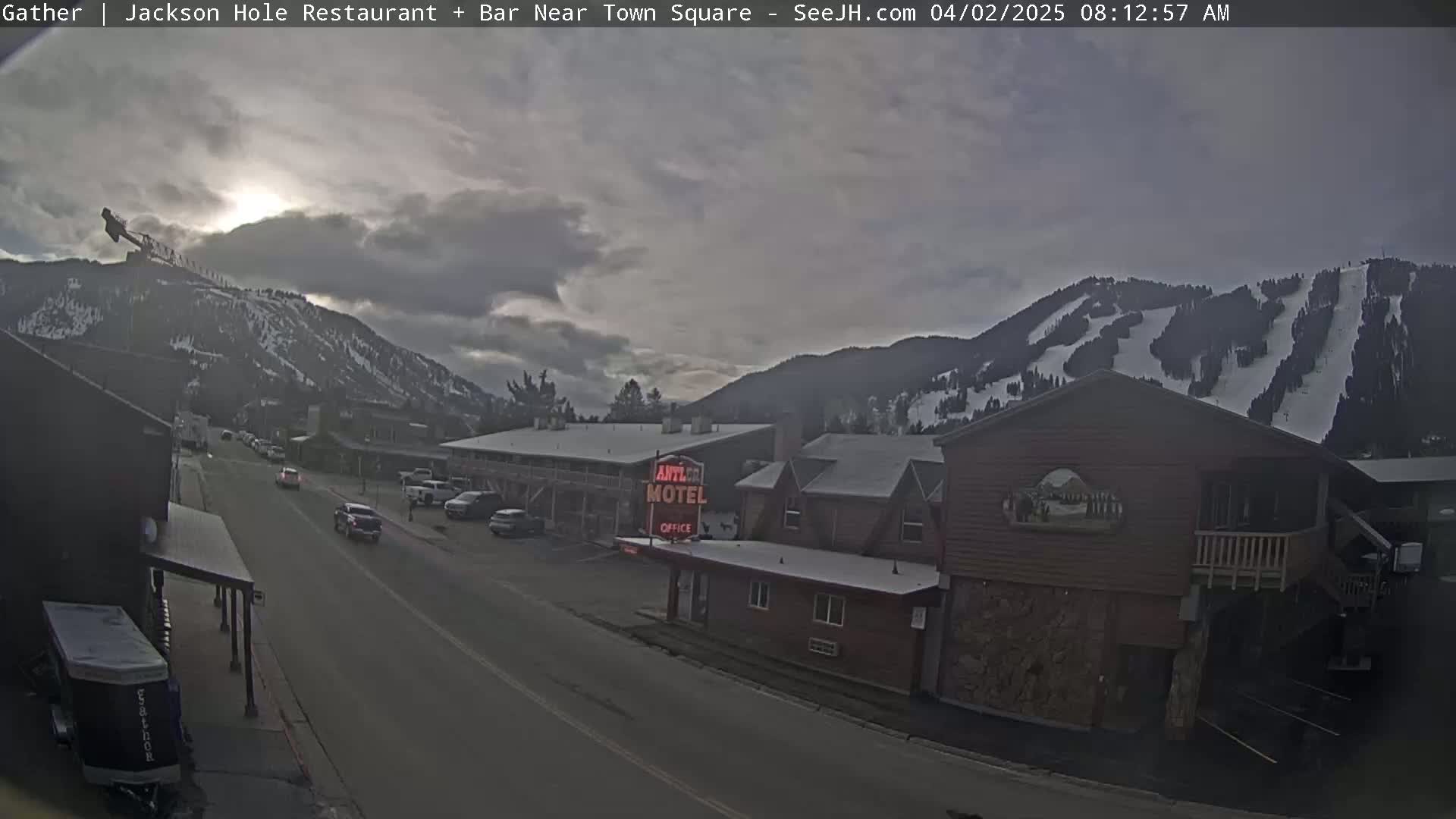 A mostly cloudy morning scene shows a street with cars parked and driving, buildings including a motel, and snow-covered mountains in the background.