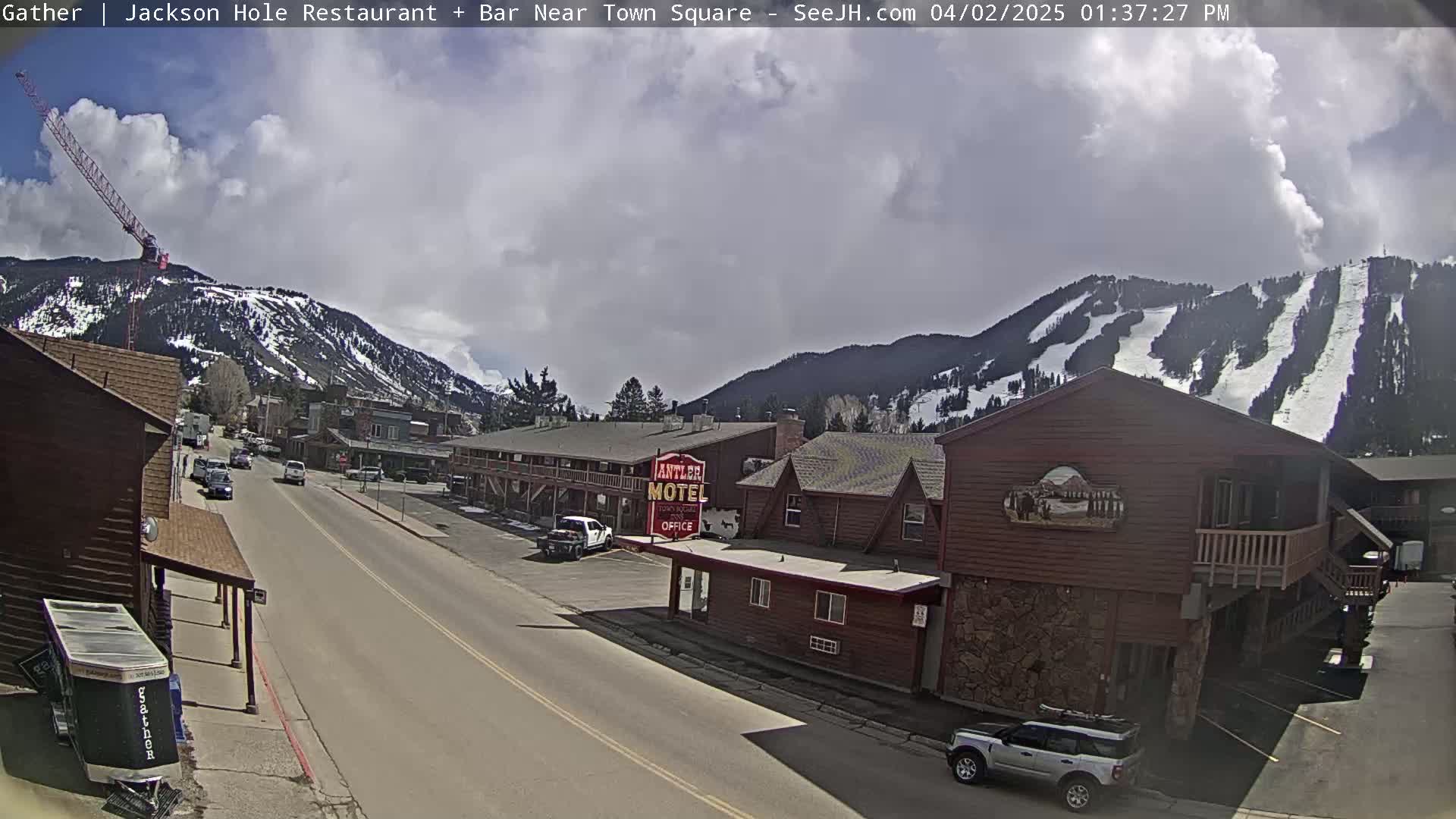 A partly cloudy day reveals a street scene in a mountain town, with cars driving on a road past a motel and other buildings, a ski slope visible on the mountainside in the background, and a construction crane in the distance.