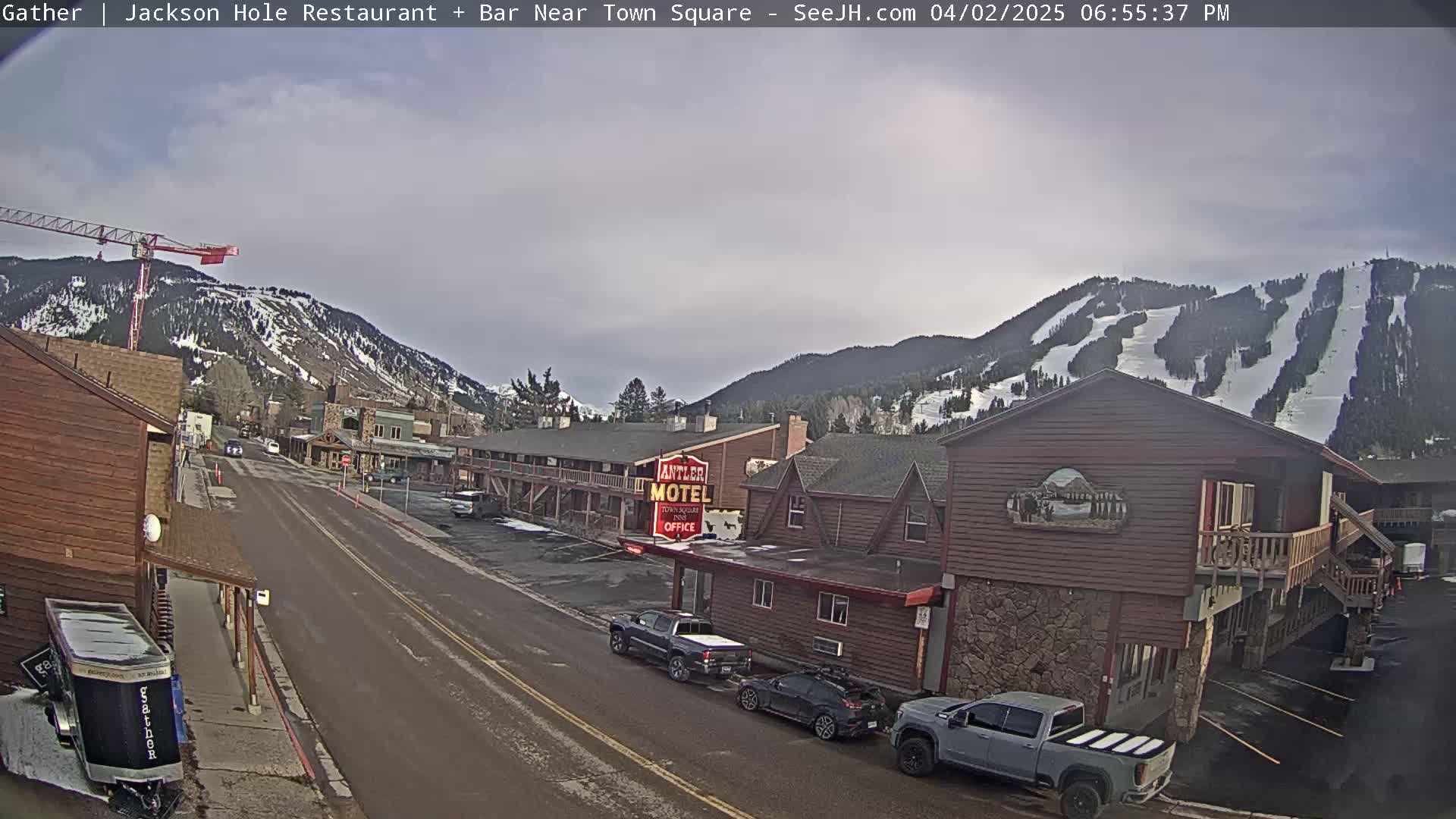 A mostly cloudy day reveals a street scene in a mountain town with parked cars, a motel, and a construction crane, backed by snow-covered hills with ski runs.