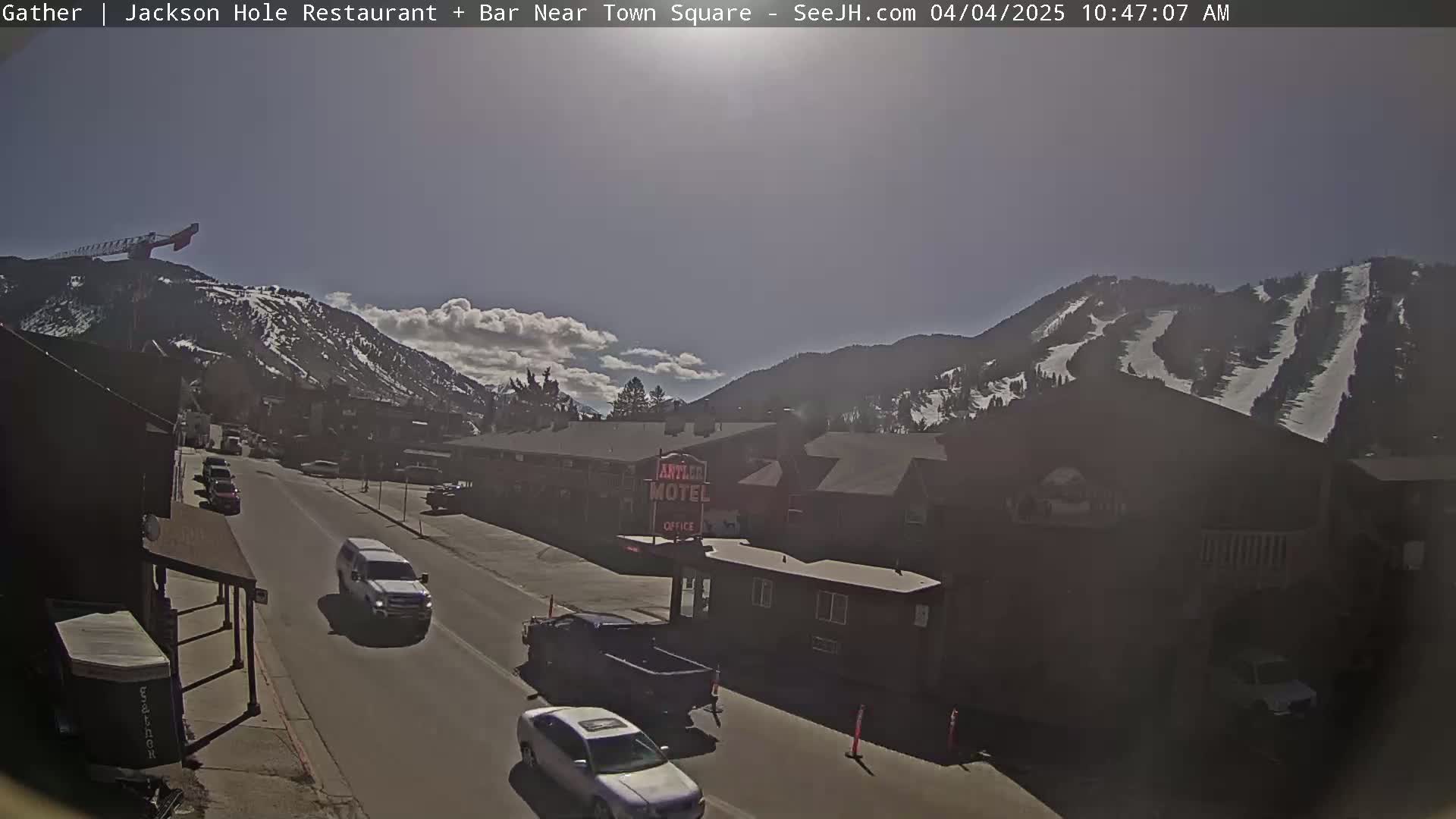A sunny day reveals a street scene with vehicles parked and driving past buildings, including a motel, against a backdrop of snow-capped mountains with visible ski slopes.