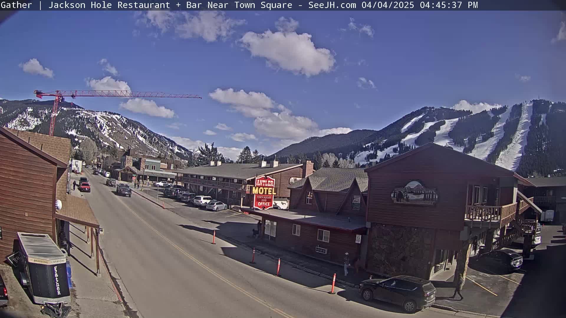 A mostly sunny day shows a street scene in a town nestled at the base of snow-covered mountains, with a construction crane visible in the distance.