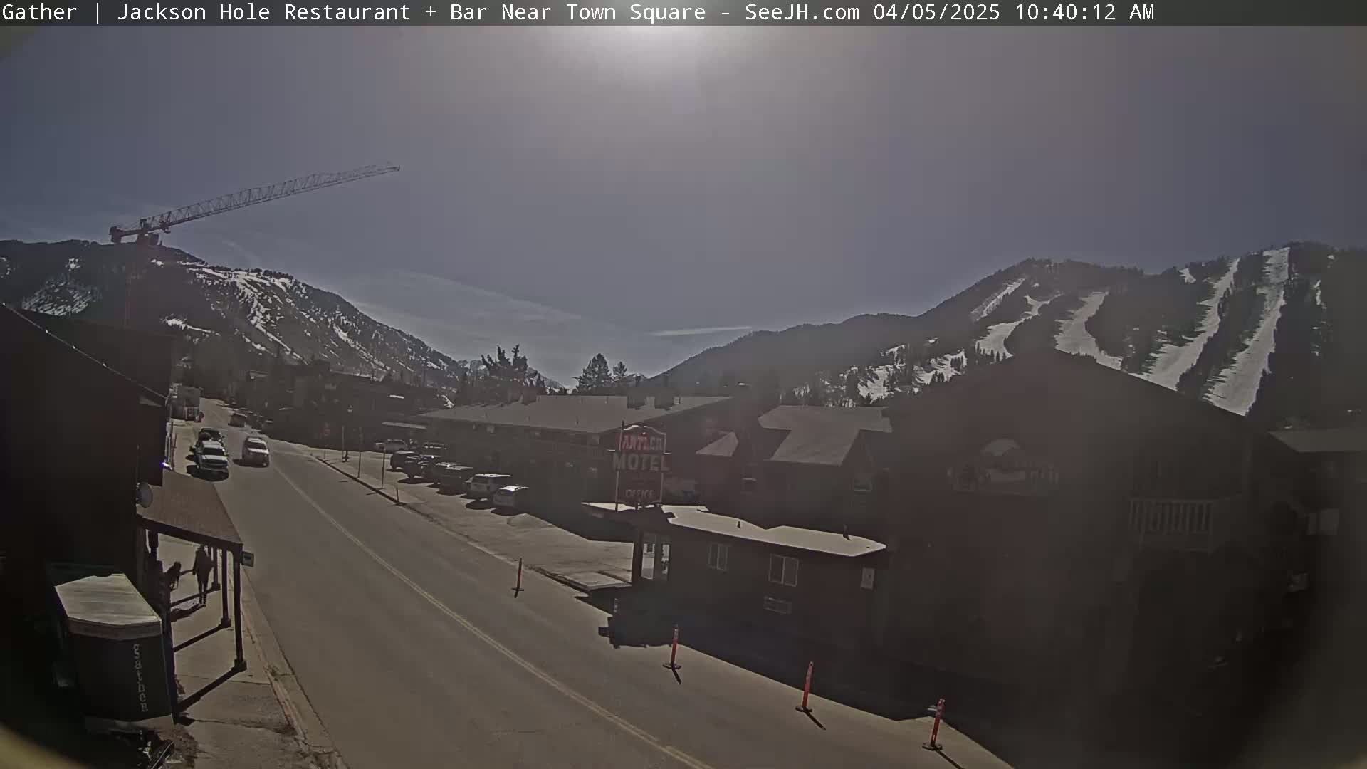 A sunny day reveals a street scene with a few cars, a motel, and a construction crane overlooking snow-covered mountains in the background.